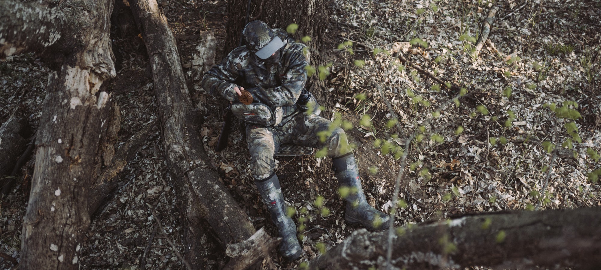Blake Wollerman sitting under a tree grabbing a pot call out of the all-new Turkey Tool Belt wearing the Equinox Guard System in Optifade Timber | SITKA Gear
