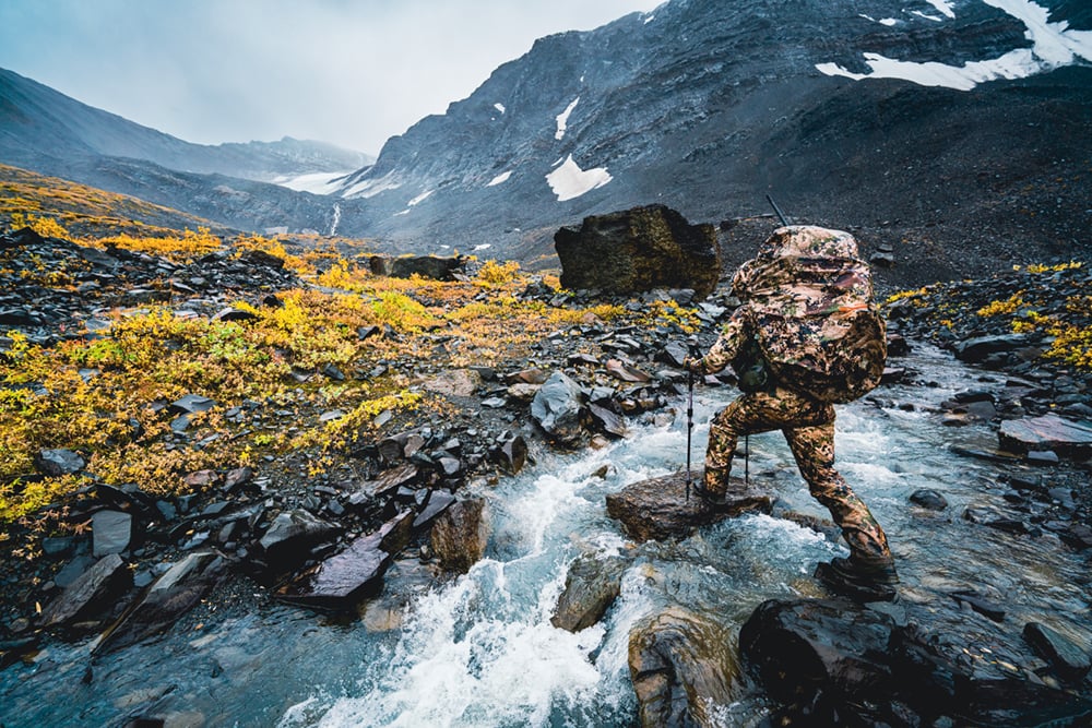 Man crossing raging river in the Dew Point Collection in Optifade Sublapline | SITKA Gear