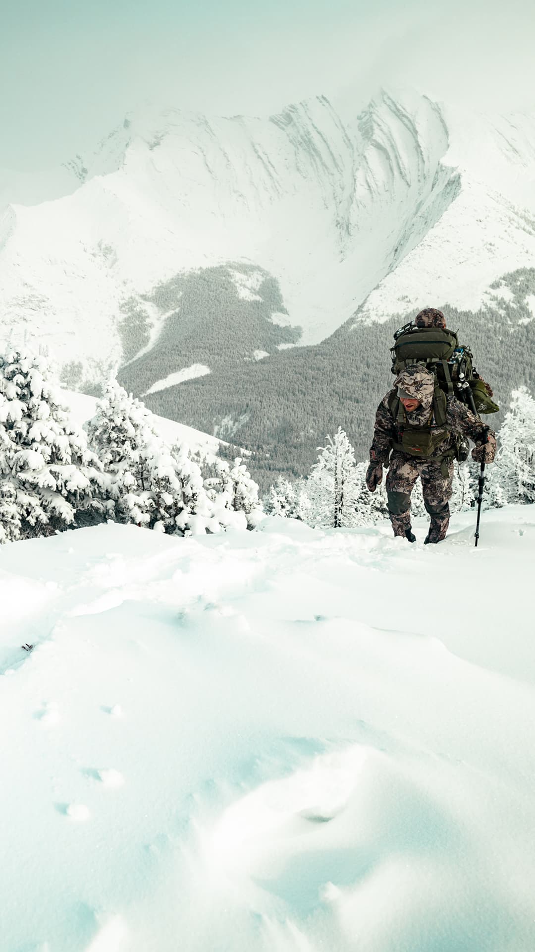 A man climbing in the snowy high alpine with a hunting pack and trekking poles.