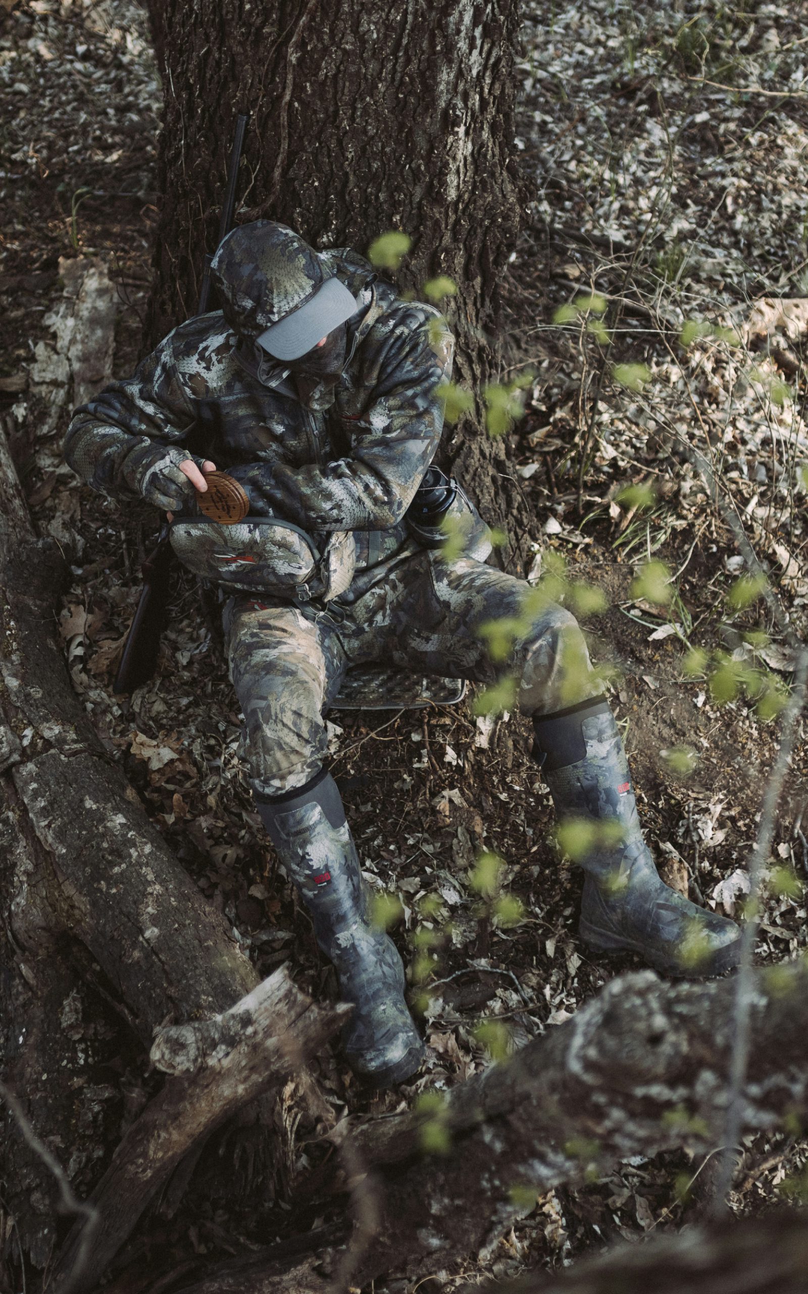Blake Wollerman sitting under a tree grabbing a pot call out of the all-new Turkey Tool Belt wearing the Equinox Guard System in Optifade Timber | SITKA Gear