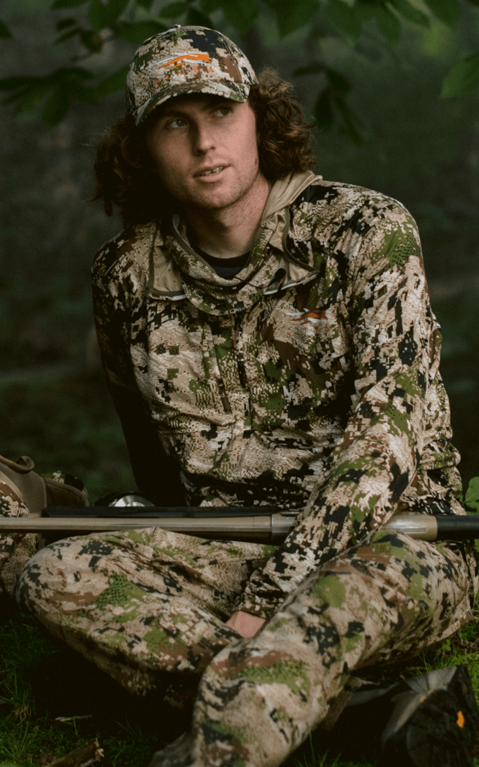 Beau Brookes sitting on grass holding a shotgun with the Turkey Tool Belt in Optifade Subalpine | SITKA Gear