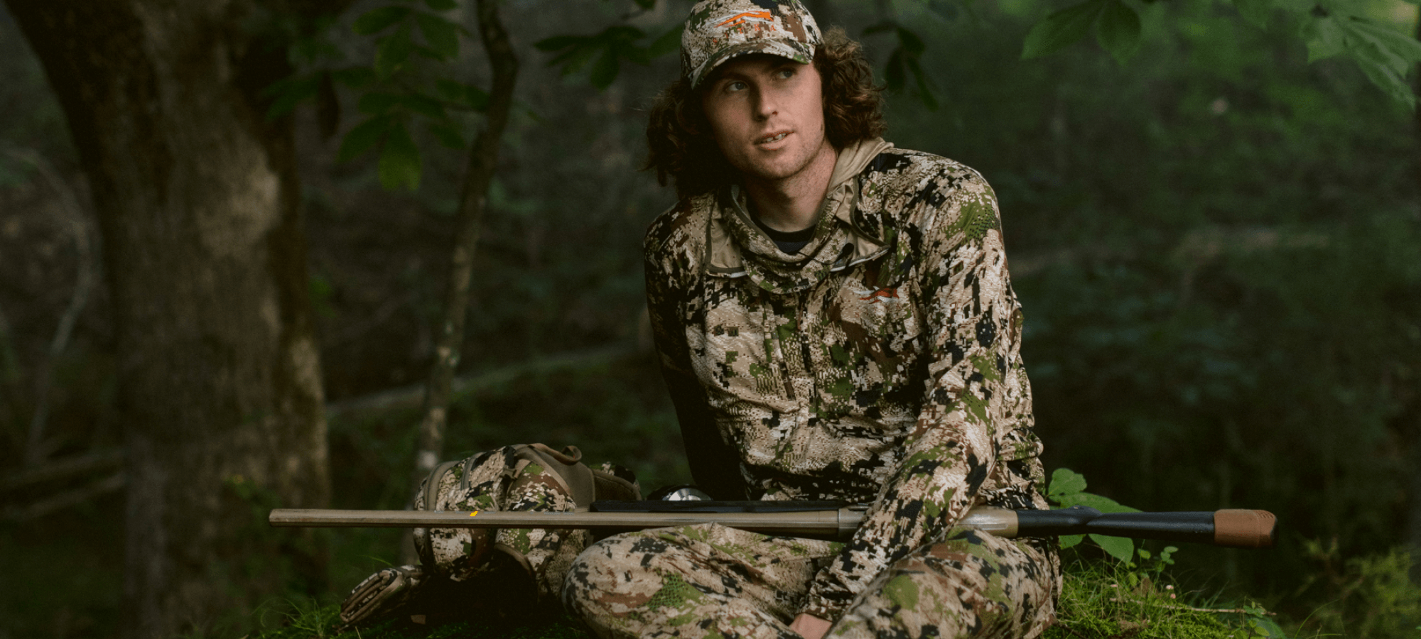 Beau Brookes sitting on grass holding a shotgun with the Turkey Tool Belt in Optifade Subalpine | SITKA Gear