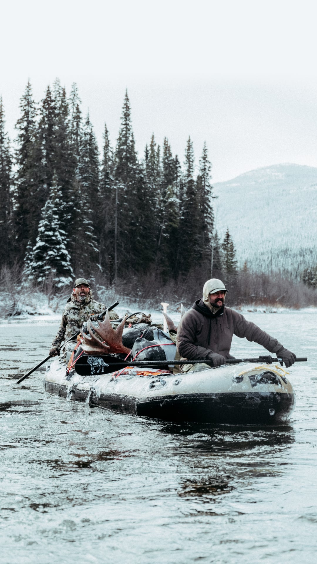 Two men bundled up on a raft packed with gear and deer floating down a cold river scene in winter.